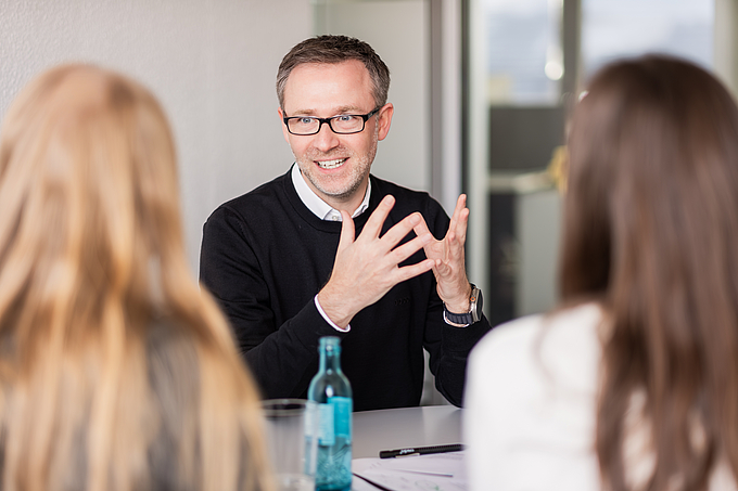Marketingexperte Björn spricht in einem modernen Besprechungsraum mit zwei Kolleginnen über eine Kommunikationsstrategie. Während er gestikulierend seine Ideen erklärt, hören die Teilnehmerinnen aufmerksam zu.