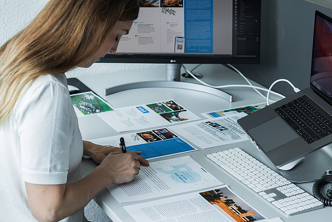 Eine Frau sitzt an ihrem Schreibtisch und schreibt. Um sie herum liegen ausgedruckte Seiten von einem Mitarbeitermagazin. das Magazin ist auch auf dem Bildschirm hinter ihr zu sehen.