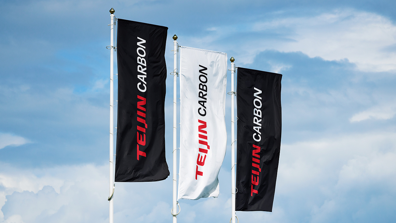 Drei wehende Flaggen, zwei schwarze und eine weiße, mit dem weißen und roten Teijin Carbon Logo und Schriftzug.