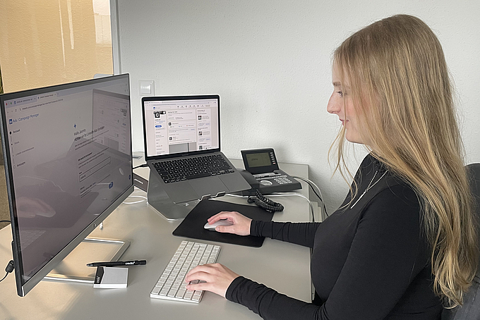 Eine Frau sitz an ihrem Schreibtisch, vor ihr stehen ein Laptop und ein Bildschirm.