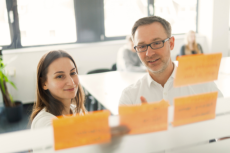 Zwei Personen arbeiten gemeinsam an einer Glaswand mit orangefarbenen Haftnotizen. Der Mann zeigt auf eine Notiz, die Frau lächelt, beide wirken konzentriert und im Austausch, im Hintergrund unscharf ein Besprechungsraum mit weiteren Personen.