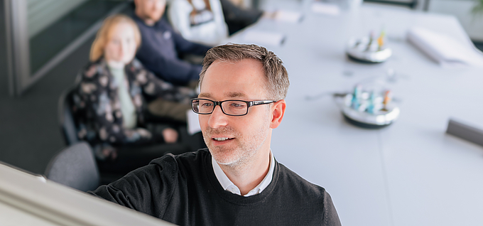 Björn präsentiert Inhalte an einem Flipchart während eines Strategieworkshops, im Hintergrund sitzt ein Team an einem Konferenztisch und hört aufmerksamzu.