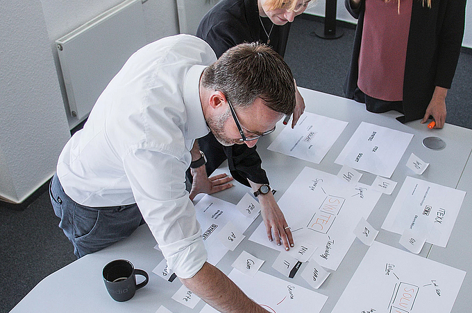 Drei Personen arbeiten gemeinsam an einem Tisch an Konzeptpapieren und Ideen, während sie Begriffe und Strukturen auf Blättern anordnen.